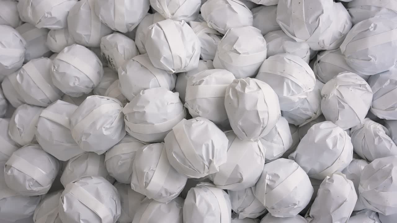 Groepen mensen verzamelen zich op een markt op een stadsplein. Ze pakken kleine papieren balletjes met lekkernijen uit. De sfeer is levendig terwijl mensen met elkaar omgaan en momenten delen.