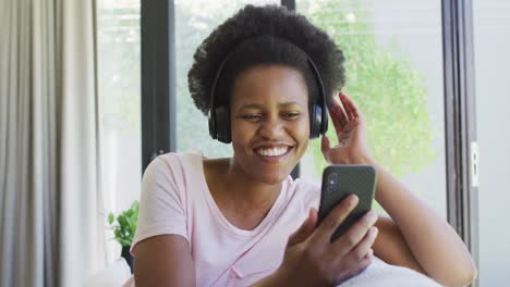 Happy-african-american-woman-wearing-headphones,-sitting-on-sofa,-using-smartphone