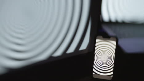 a mobile phone screen in focus, with a blurry computer and laptop screen, in a dark room with all displaying a hypnotic spiral pattern