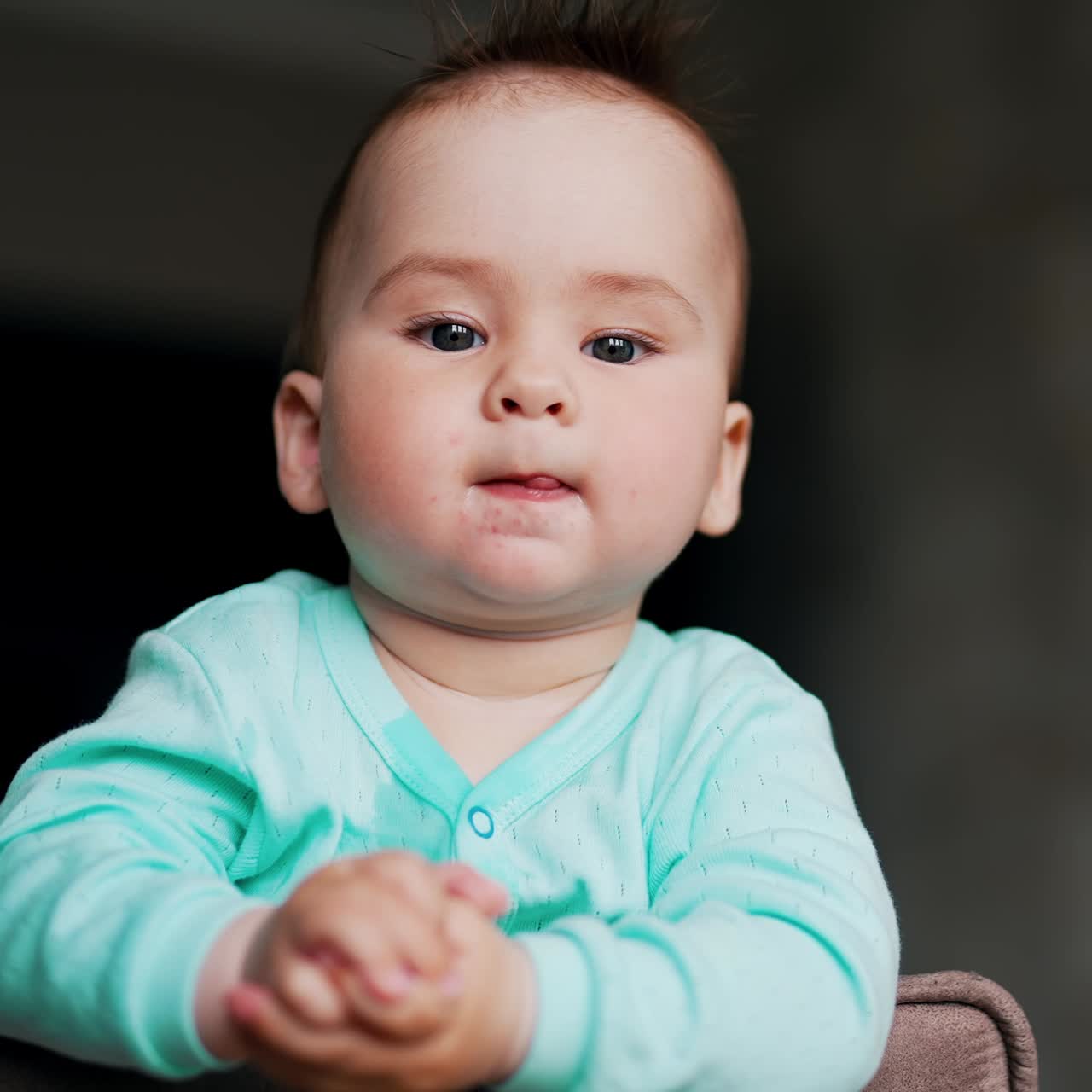Nice Caucasian boy with sticking out hair looks somewhere attentively. Sweet kid in blue shirt on a chair. Blurred dark background