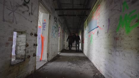 Two-men-walking-along-dark-shadowy-eerie-extended-corridor,-slow-motion