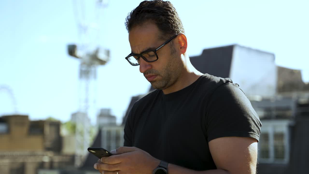 Uomo atletico europeo con gli occhiali che usa intensamente il suo smartphone all'aperto, mostrando concentrazione e tecnologia moderna.