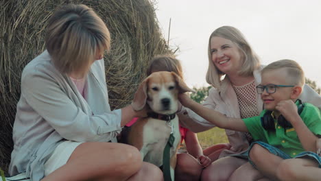 family seated outdoors affectionately touching their dog, sharing a moment of closeness near a hay bale during sunset, joyful expressions in a rural setting