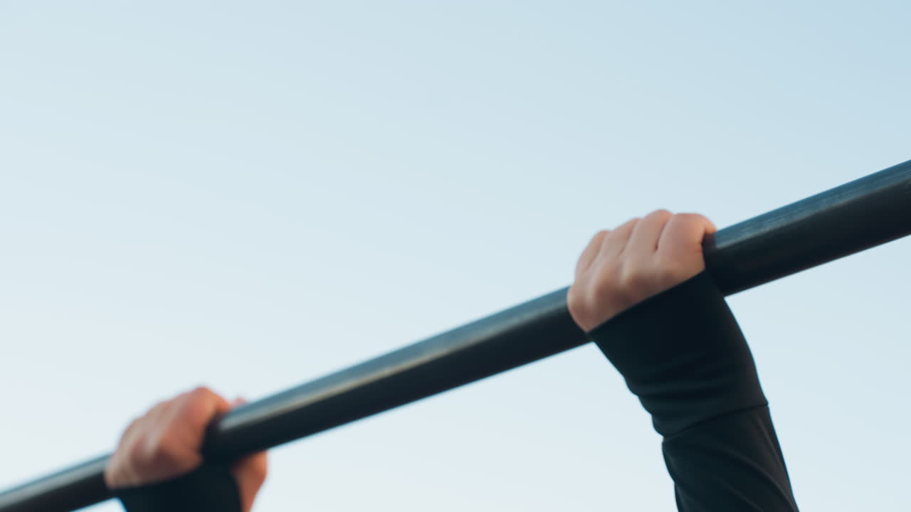 Sky view of female hands gripping horizontal bar for balance against pale blue sky conveying focus strength and stability during urban outdoor workout preparation showcasing fitness routine
