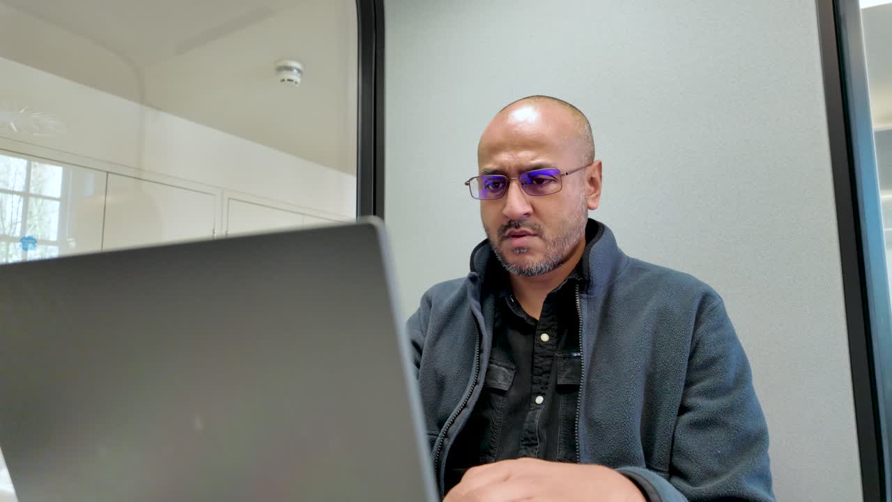 A focused South Asian man working on his laptop in a private office pod.