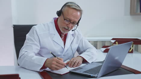 Remote medicine. Senior male specialist giving a consultation to a patient through video call. Medical specialist in headphones talking and writing treatment.