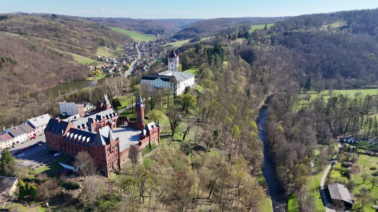 Bevægelse mod højre, mens dronen langsomt roterer og daler, afslører Hradec nad Moravicí slotkomplekset i Tjekkiet med floddal, træer og et bølgende landskab