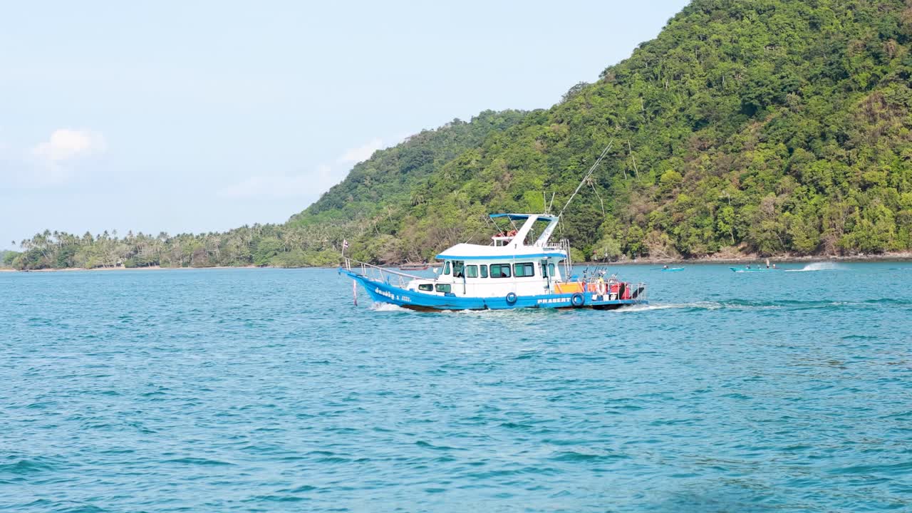 A blue fishing boat sails near lush green hills in Phuket, Thailand, under clear skies and calm seas