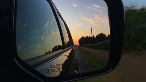looking in a car rear view mirror while driving next to corn fields in illinois at sunset.