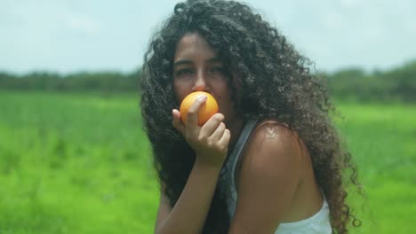 young woman with voluminous type 3c curls and warm brown skin holds a peach-like fruit close to her mouth, staring at the camera in a vivid green field under bright midday light