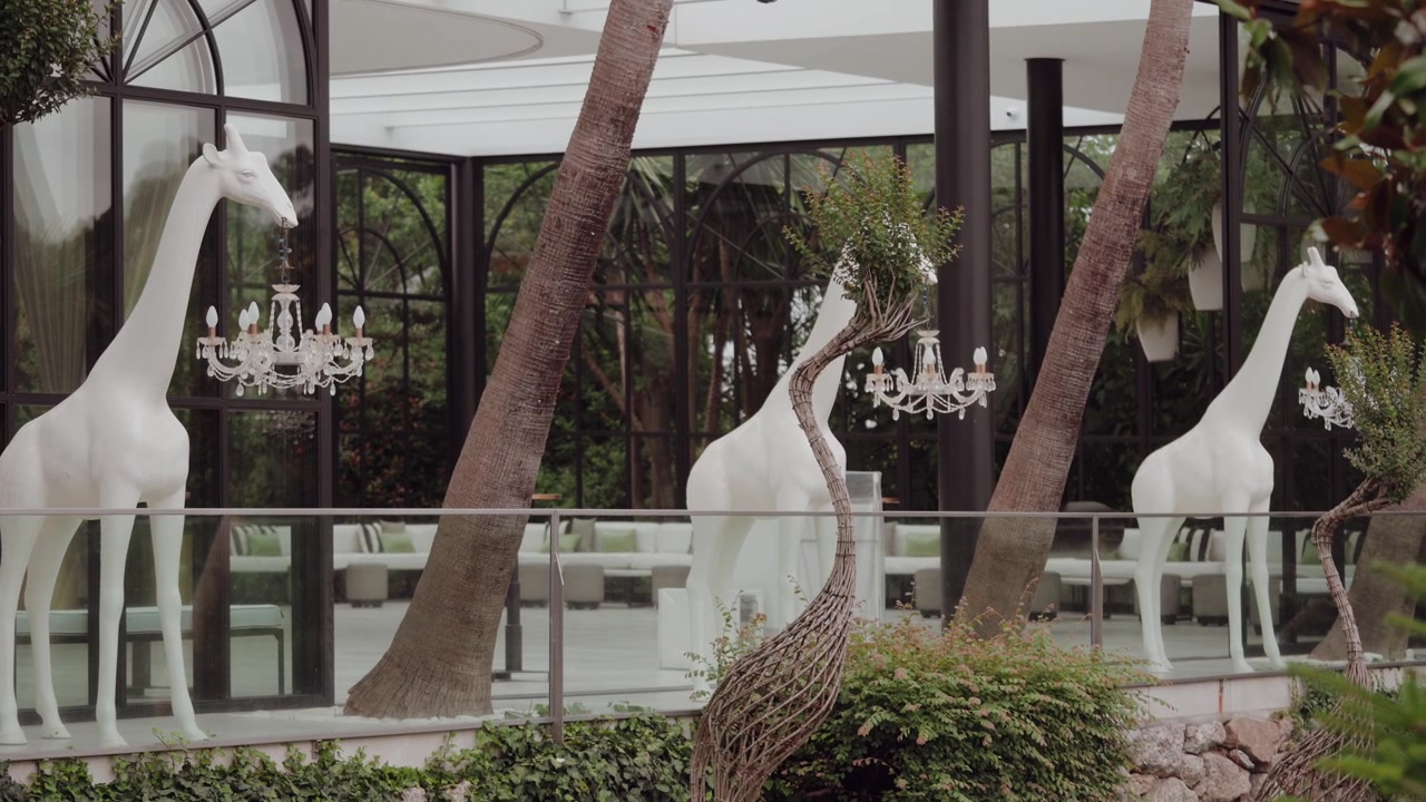 Estatuas de jirafas blancas de tamaño natural junto a troncos de palmeras y candelabros de cristal en un recinto acristalado.