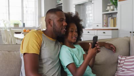 Hija-Afroamericana-Y-Su-Padre-Usando-Un-Teléfono-Inteligente-Juntos-Sentados-En-El-Sofá