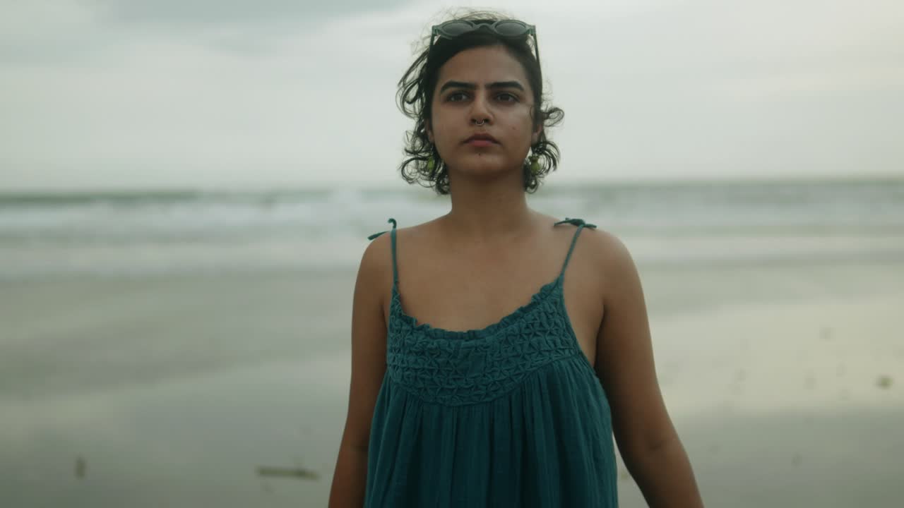 una atractiva mujer india en cámara lenta, con gafas de sol en la cabeza, mira directamente a la cámara en un día nublado en la playa
