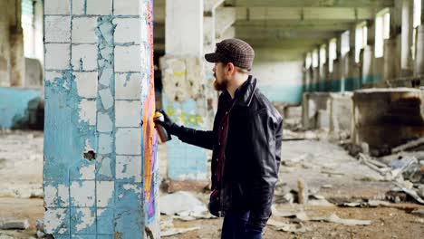 handsome bearded guy is holding aerosol paint and drawing graffiti on pillar inside spacious abandoned house. creative people, empty buildings and modern art concept.