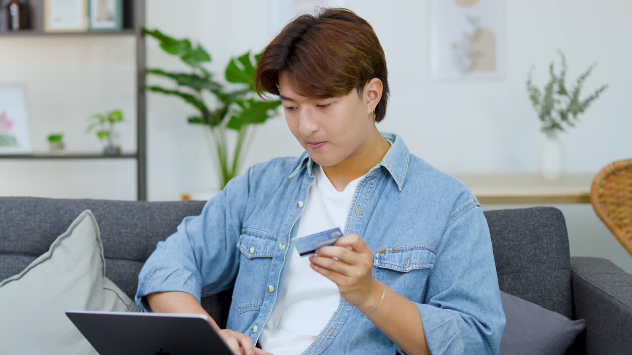 若いアジア人男性がソファに座り、ノートパソコンにクレジットカード情報を入力しています。