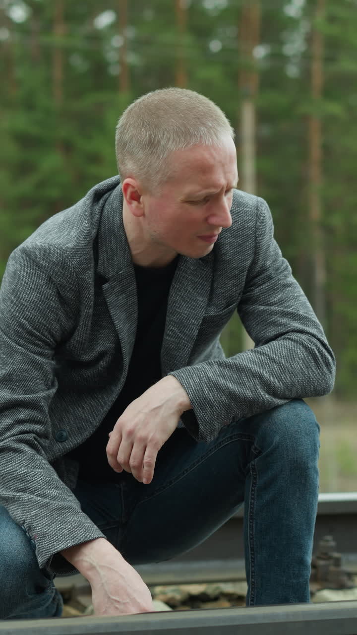 A close-up view of a man in a grey blazer and blue jeans kneeling by a railway track, examining it closely with a thoughtful expression