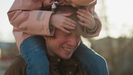 father with a warm smile as his little girl in a pink jacket playfully places both her hands and head on his, outdoors at sunset