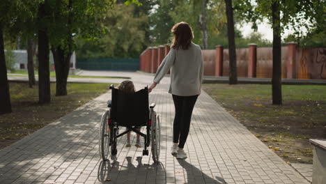 woman walks on road moving wheelchair with little daughter