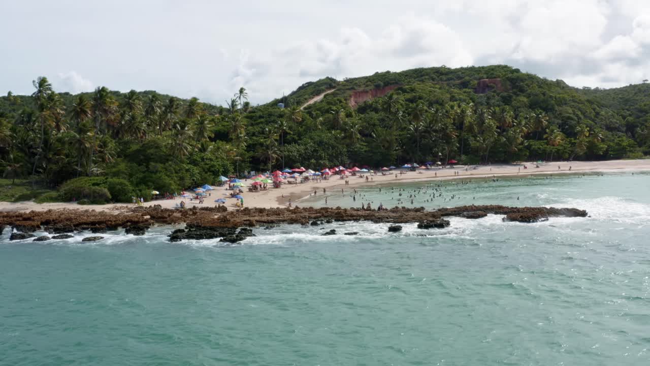 vista aérea de la popular playa tropical de coquerinhos cubierta de paraguas con turistas nadando en una piscina natural desde un arrecife que bloquea pequeñas olas en condé, paraíba, brasil