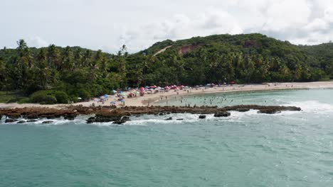 Vista-Aérea-De-Drones-De-La-Popular-Playa-Tropical-De-Coquerinhos-Cubierta-De-Sombrillas-Con-Turistas-Nadando-En-Una-Piscina-Natural-Desde-Un-Arrecife-Bloqueando-Pequeñas-Olas-En-Conde,-Paraiba,-Brasil