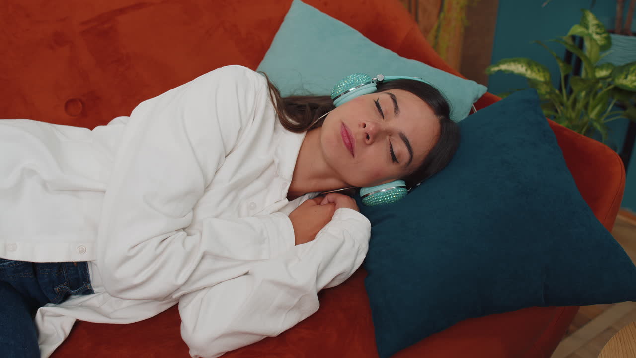 무선 헤드폰을 입은 행복한 젊은 여성이 집에서 소파에 누워서 좋아하는 음악을 듣고 휴식을 취하고 있습니다.