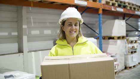 feliz empleada sosteniendo una caja de cartón en el almacén y mirando la cámara