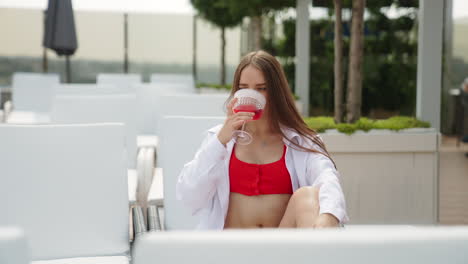 having cocktails at hot summer day near the pool. young attractive brunette in red swimsuit drinks red beverage from a wineglass.