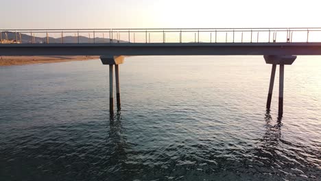 side view of a jetty at the beach, flying alongside the metallic structure reflecting in water in the gentle light of the setting sun