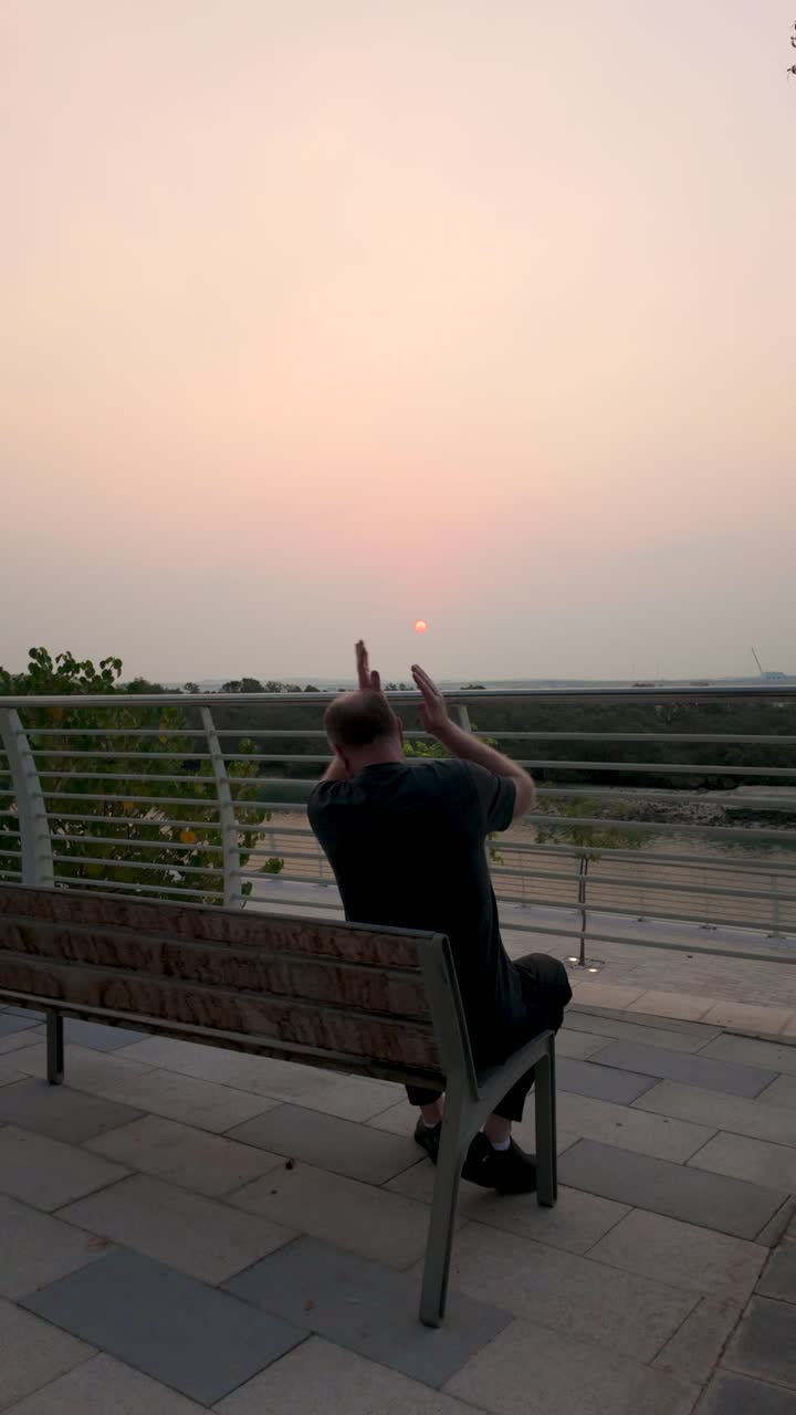 A man sits on a park bench watching a serene sunrise. Filmed in vertical video format, perfect for social media. Calm morning atmosphere, slightly bothered by insects