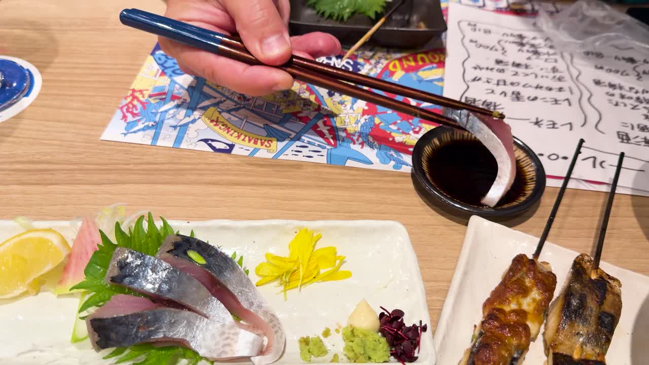 A person uses chopsticks to dip fresh mackerel sashimi into soy sauce at an izakaya