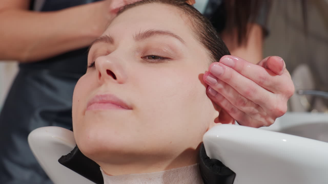 미용실 세면대에서 딥 컨디셔닝 마스크와 두피 마사지를 받고 있는 백인 모델, 스타일리스트가 빗으로 크림을 바르고 목 보호대가 제자리에 있으며, 헤어 트리트먼트가 수분과 윤기를 회복시켜 주고 편안해 보입니다.