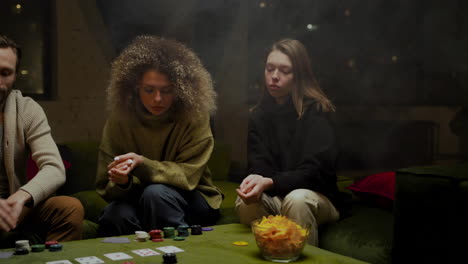 group of friends playing poker sitting on the couch while eating potato chips in a big glass bowl