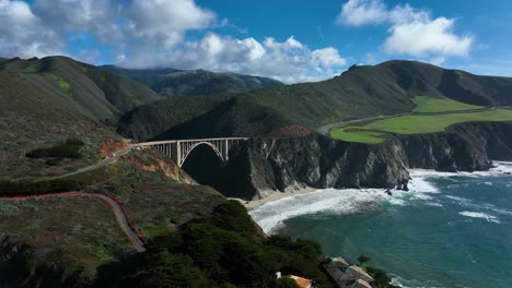 Filmische-Ansicht-Der-Bixby-Bridge-Mit-Den-Grünen-Santa-Lucia-bergen-Im-Hintergrund,-Während-Unten-Die-Wellen-Krachen,-Kalifornien