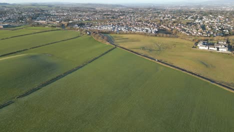 Vista-Aérea-De-Un-Campo-Verde-Con-Una-Carretera-Y-Una-Gran-Ciudad-Al-Fondo