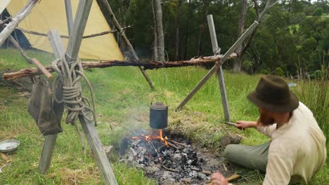 bushman lying by a campfire cooking food on a billy by a historical hut