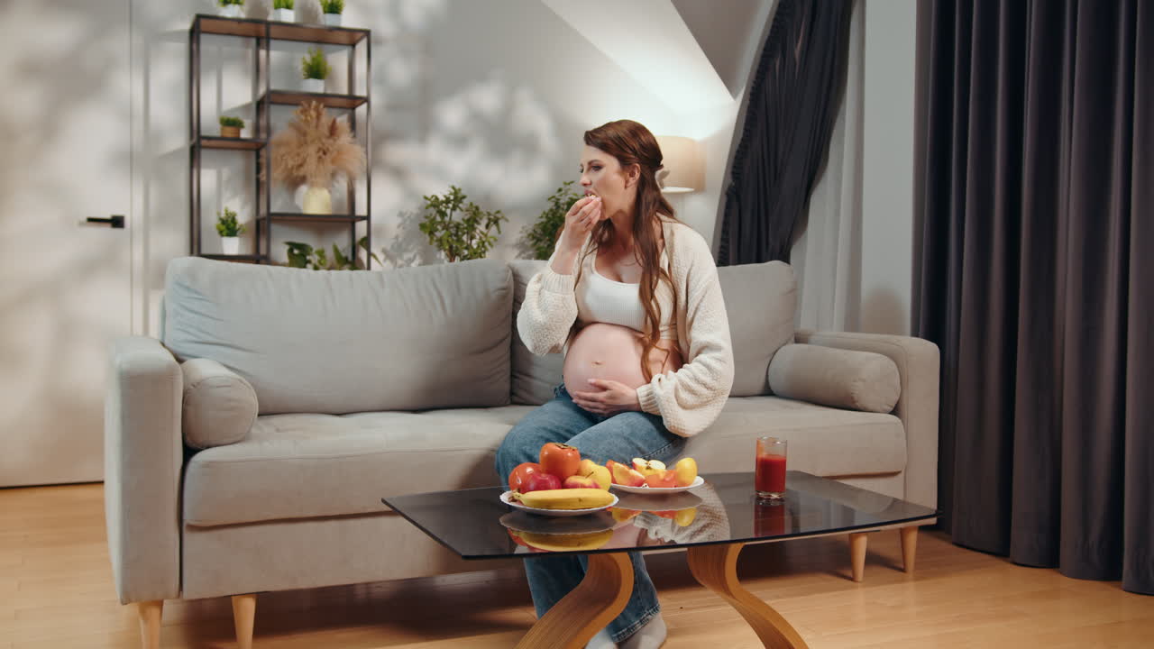 Pregnant woman sits on sofa eating fruit and holding her belly healthy food on coffee table pregnant