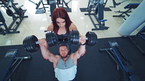 portrait of athletic man with muscular body lifting dumbbells in gym. strong man lifts heavy dumbbells with the help of beautiful red-haired woman in sport club indoors. top view.