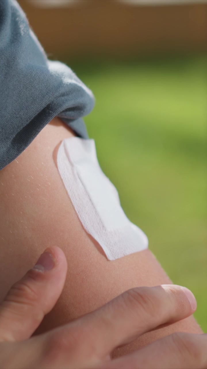 Primo piano di una persona che applica un cerotto adesivo al braccio con arrossamento visibile. La mano tiene del nastro medico bianco, rivelando la texture della pelle e piccole abrasioni, in una luce naturale soffusa contro uno sfondo sfocato.