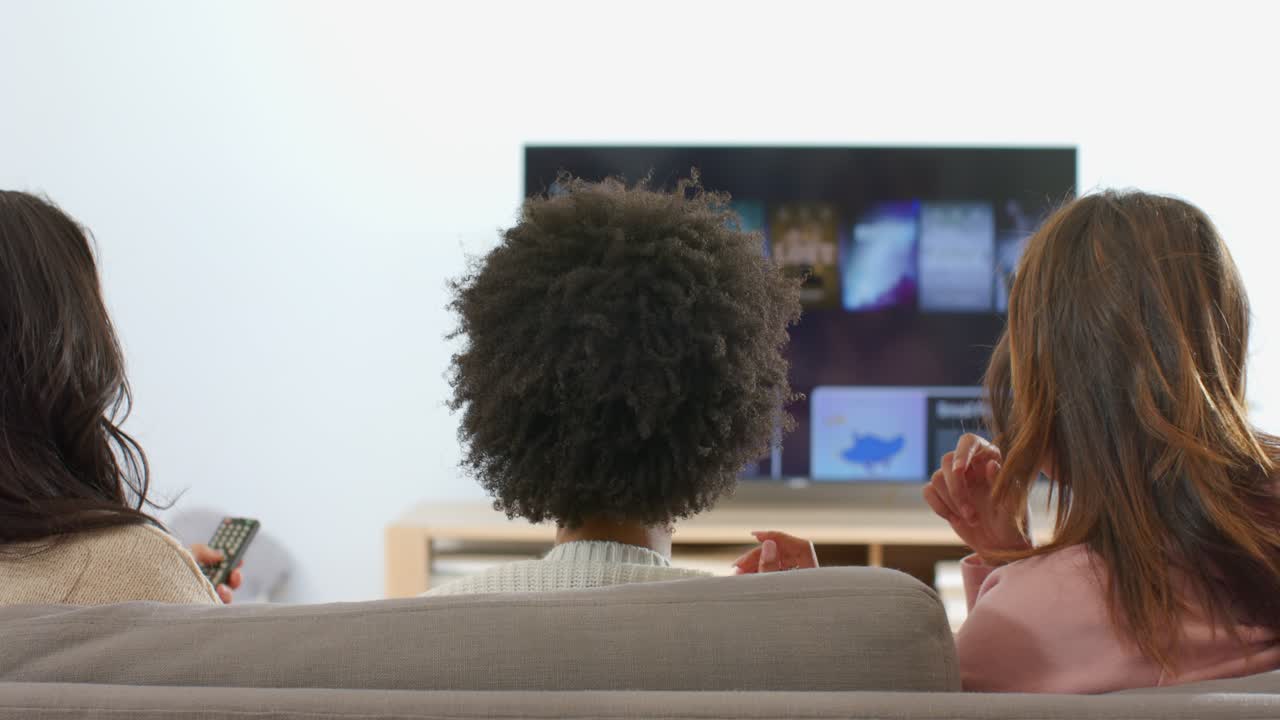 Diverse amiche che indicano il menu della TV, scorrendo con il telecomando e condividendo popcorn per scegliere un programma sul divano
