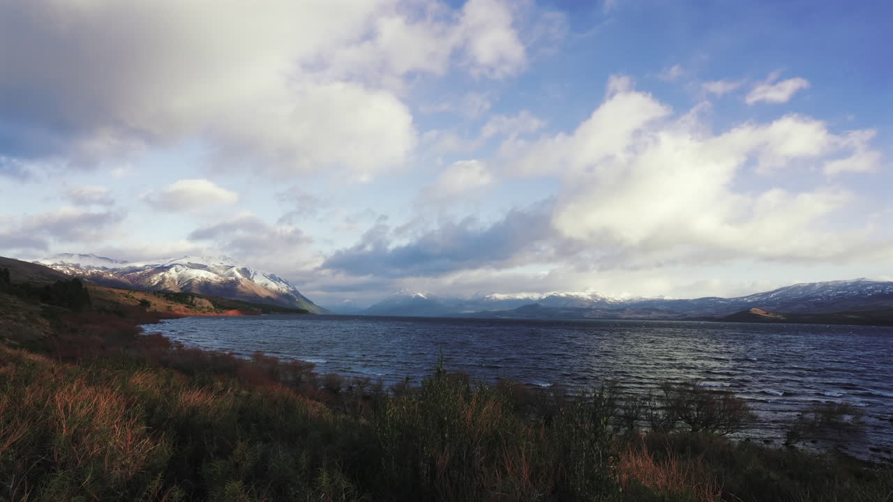 Timelapse footage of Lago Huechulafquen in Neuquén, Argentina, showing dynamic clouds and rippling waves under changing light with snow-capped peaks in the distance, static camera