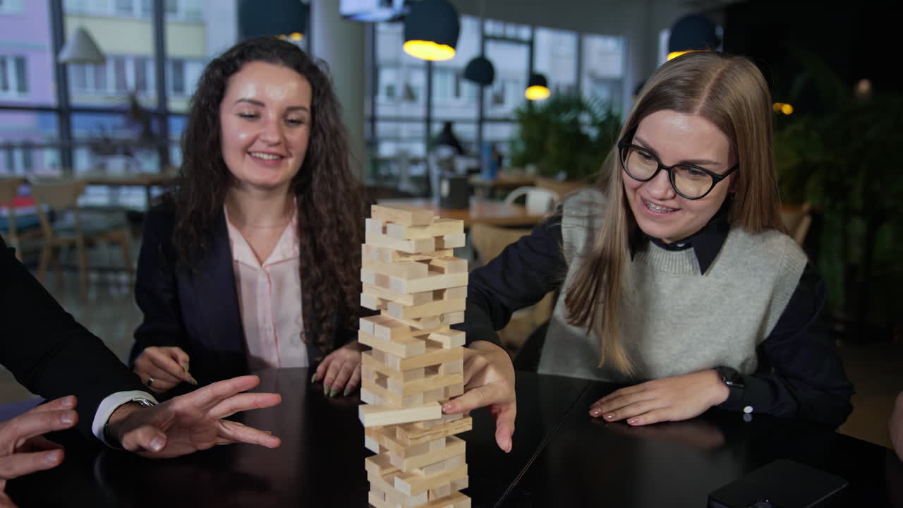 Company of friends playing jenga and having fun. One of the girls carefully pushes the brick in the pile of jenga. Leisure time for adults.