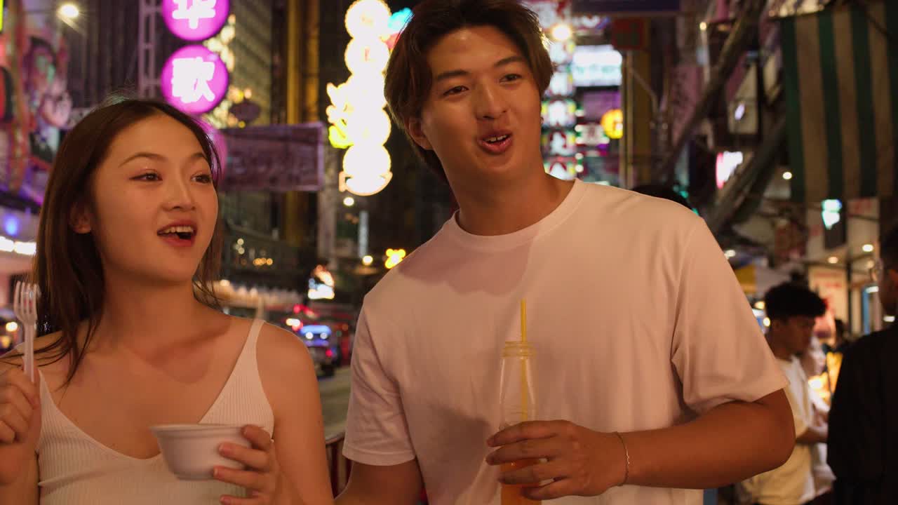 Asian couple eating, drinking, and walking through neon-lit Chinatown street at night, handheld camera