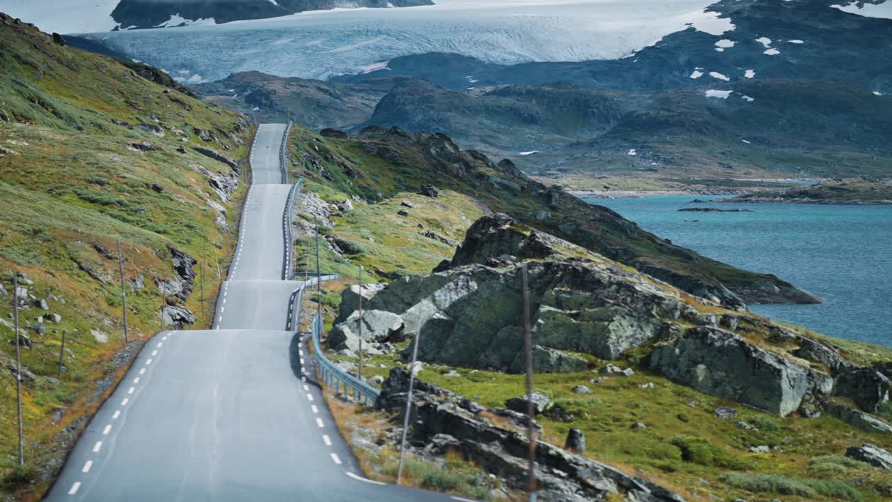 A narrow Sognefjellsvegen mountain road winds through a rocky landscape a glacier dominating the background. Parallax shot.