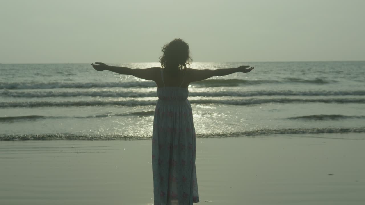 한 여성의 실루이 인도 열대 해변에 서 있고, 팔을 펴고, 태양이 그녀를 비추고 있습니다.