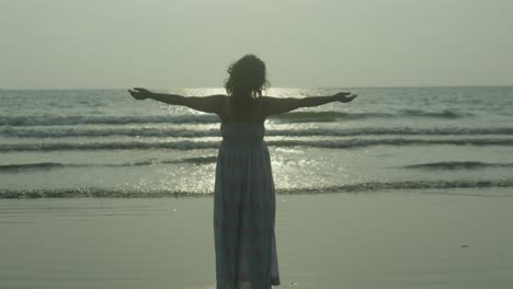 una silueta de mujer de pie en la playa tropical india, brazos extendidos, con el sol brillando sobre ella