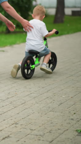a caring mother guides her little boy by pushing him gently as he starts riding his bicycle along an interlocked pathway, he begins to ride faster, another child rides in the background