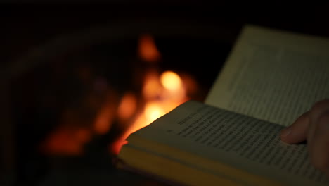 a man reading a book close to an open fire in a fireplace in a dark room