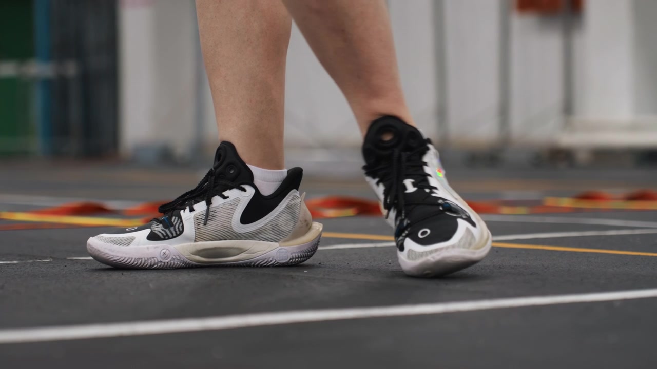 Approccio al campo da badminton che mostra passi dinamici, scarpe da ginnastica bianche e nere che eseguono un movimento potente preparandosi per un salto e uno smash su una superficie interna, con intensità, concentrazione e un posizionamento energico del piede.