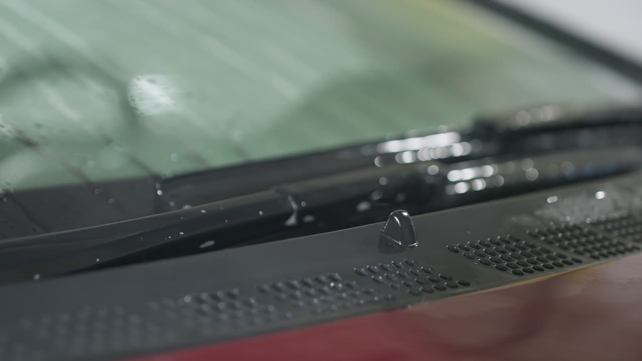 close up of black wiper blades resting on moist windshield and bonnet surface of red car with visible water droplets and reflections during cleaning process inside garage under bright lighting
