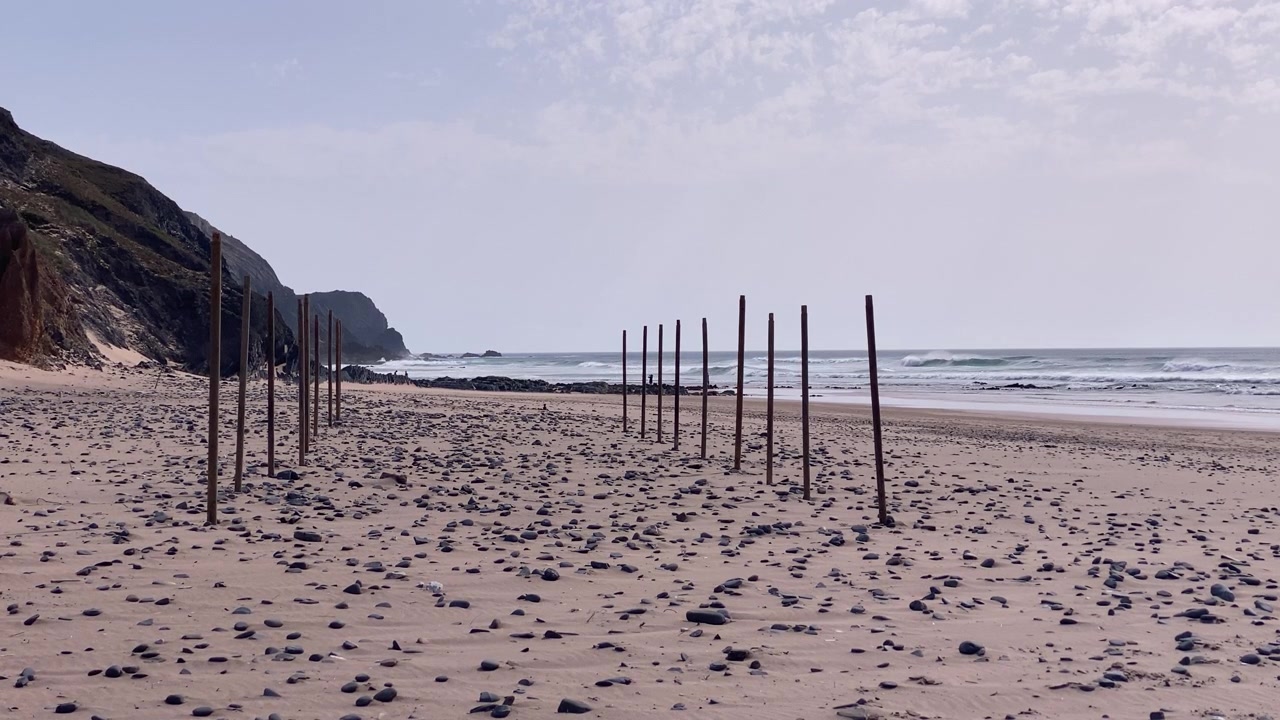 Sandstrand med rader av trestolper, småstein og eroderende kystklipper under en lys himmel i Portugal